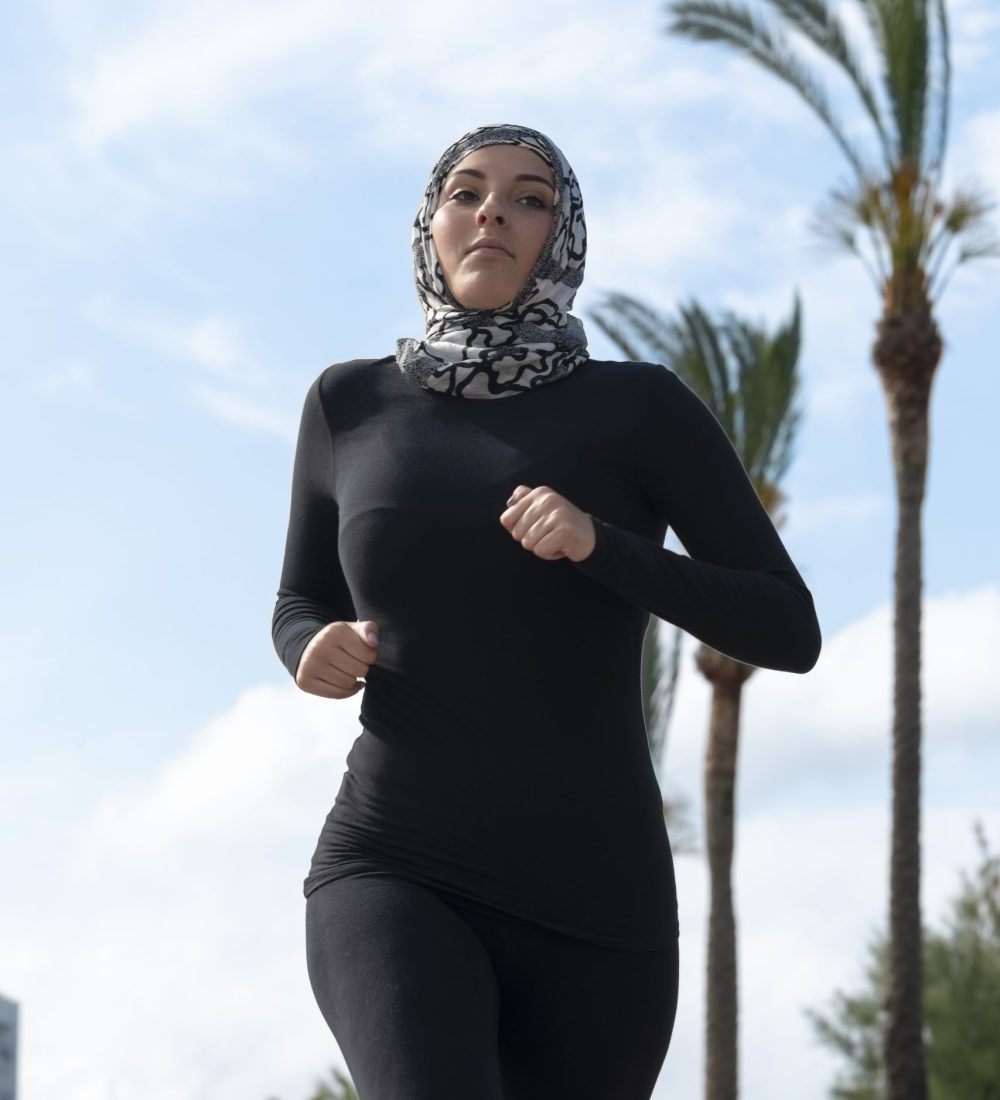 Young woman wearing a black and white headscarf, black long sleeve sports shirt and black tights running on an out of focus background. Running and diversity concept.